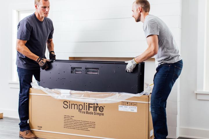 Homeowner and helper pulling electric fireplace out of shipping box in front of installation site.