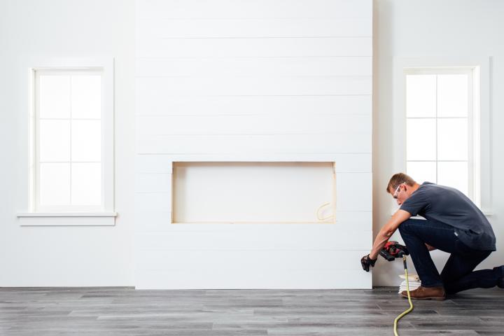 Homeowner with nail gun kneeling to install final piece of shiplap on electric fireplace wood chase.