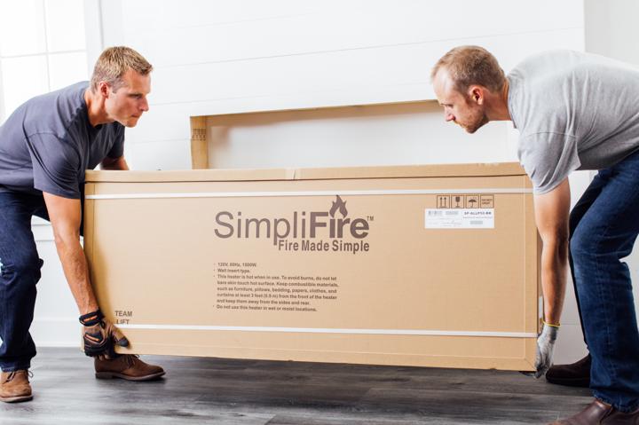 Homeowner and helper placing boxed electric fireplace on floor in front of installation site.