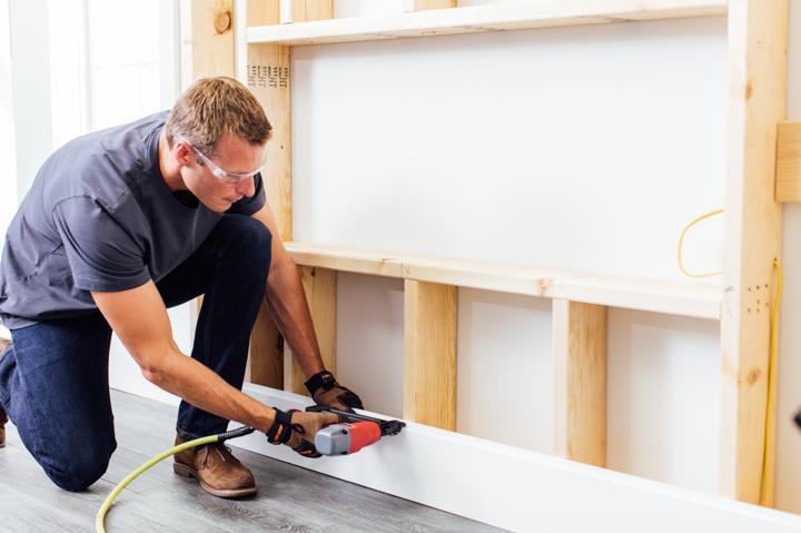 Homeowner with nail gun kneeling to install bottom piece of white shiplap on electric fireplace wood chase.