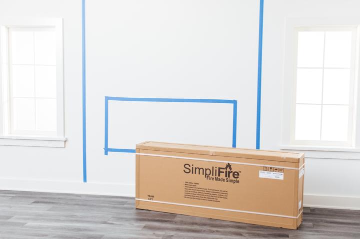 Shipping box containing electric fireplace in front of installation wall marked with placement area in blue tape.